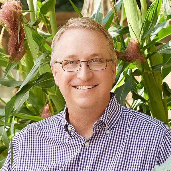 a man wearing glasses standing in front of a plant.