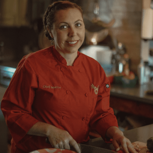a woman in a red chef's coat preparing food in a kitchen.