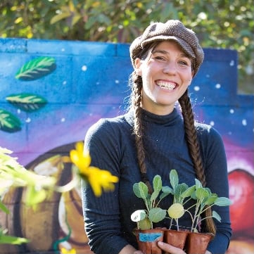 a smiling woman holding a potted plant in front of a mural.